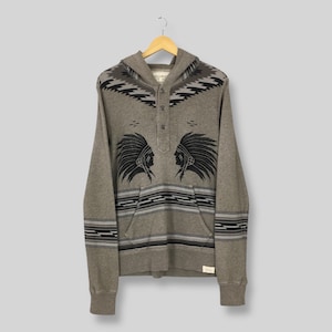 May include: A grey hoodie with a hood and a kangaroo pocket. The hoodie features a Native American design with two profiles wearing headdresses. The design is in black and grey. The hoodie has a button-up neckline and striped patterns.