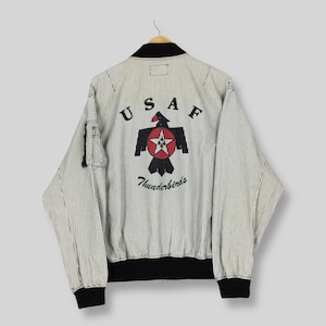 Könnte beinhalten: Vintage Bomberjacke mit schwarz-weißem Streifenmuster. Auf der Rückseite befindet sich ein rotes, schwarzes und weißes Thunderbird-Logo mit "USAF" darüber und "Thunderbirds" darunter. Die Jacke hat schwarze Bündchen, Kragen und Saum.