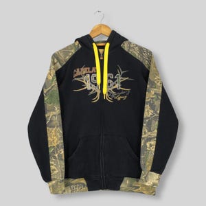 Puede incluir: Sudadera negra con cremallera, cordón amarillo y mangas de camuflaje. La parte delantera presenta el texto "Cabela's 1961 Legendary Legacy" en marrón y dorado. La sudadera tiene un bolsillo delantero y está colgada de una percha de madera.