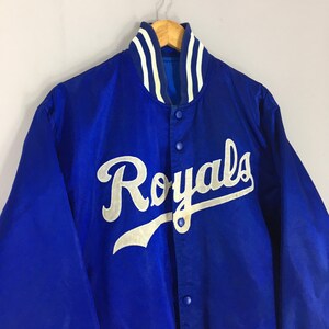Pu&ograve; includere: Giacca in raso blu reale con la scritta "Royals" in corsivo bianco sul petto. Il colletto e i polsini hanno strisce bianche e blu. La giacca &egrave; abbottonata e appesa a una gruccia di legno.