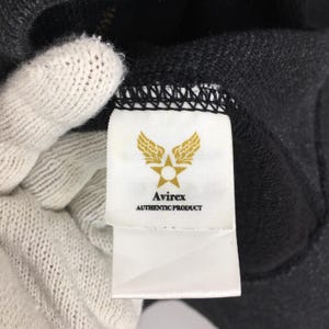 Pu&ograve; includere: Primo piano di un'etichetta bianca con il logo Avirex e la scritta "AUTHENTIC PRODUCT" su un capo grigio scuro. Il logo presenta una stella all'interno di un cerchio, affiancata da ali stilizzate. Il capo sembra essere una felpa con cappuccio.