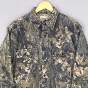 May include: Camouflage button-down shirt with two chest pockets. The shirt features a woodland camouflage pattern in shades of green, brown, and gray. The brand "NORTHWEST" is visible on the label. The shirt is hanging on a wooden hanger.