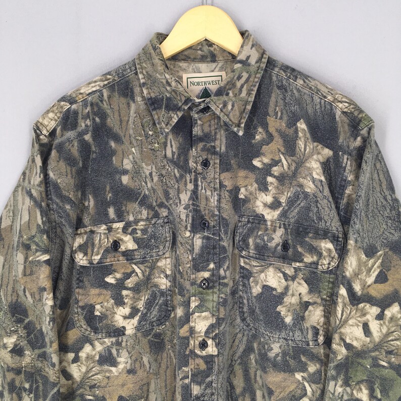 May include: Camouflage button-down shirt with two chest pockets. The shirt features a woodland camouflage pattern in shades of green, brown, and gray. The brand "NORTHWEST" is visible on the label. The shirt is hanging on a wooden hanger.