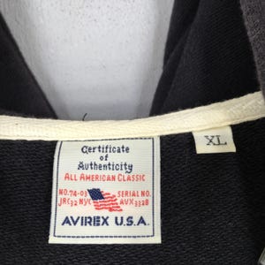 Pu&ograve; includere: Primo piano di una felpa con cappuccio nera con coulisse bianca ed etichetta con la scritta "Certificate of Authenticity, All American Classic, AVIREX U.S.A.". L'etichetta presenta anche una piccola bandiera americana e la taglia "XL".