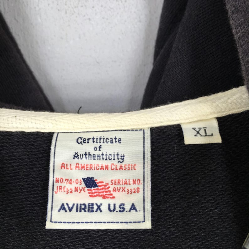Pu&ograve; includere: Primo piano di una felpa con cappuccio nera con coulisse bianca ed etichetta con la scritta "Certificate of Authenticity, All American Classic, AVIREX U.S.A.". L'etichetta presenta anche una piccola bandiera americana e la taglia "XL".