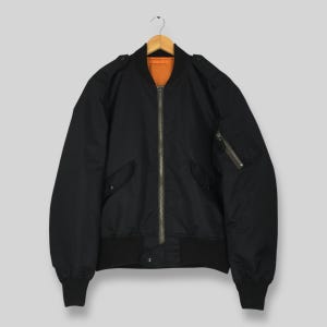 May include: A black bomber jacket with a zipper closure, ribbed cuffs, and a ribbed hem. The jacket has two front pockets and a zippered pocket on the left sleeve. The interior lining is orange. The jacket is hanging on a wooden hanger.