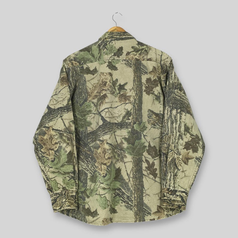 Puede incluir: Camisa de manga larga con un estampado de camuflaje en tonos verdes, marrones y beige, que imita una escena forestal. La camisa est&aacute; colgada de una percha de madera, mostrando la parte trasera de la prenda.