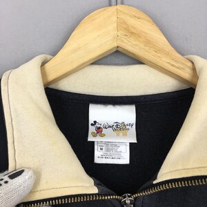 May include: Black zip-up jacket with a cream-colored collar and a wooden hanger. The tag features the Walt Disney World logo with Mickey Mouse. The jacket has a zipper and a size M label.