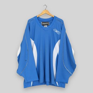 May include: A blue and white Reebok hockey jersey hanging on a wooden hanger. The jersey has a V-neck and white accents on the sleeves and sides. The Reebok logo is visible on the chest.