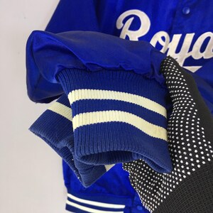 Pu&ograve; includere: Primo piano di una manica di giacca da baseball in raso blu. Il polsino della manica &egrave; blu con strisce bianche. La giacca ha la scritta "Royals" in bianco sul retro. La giacca &egrave; uno stile classico.