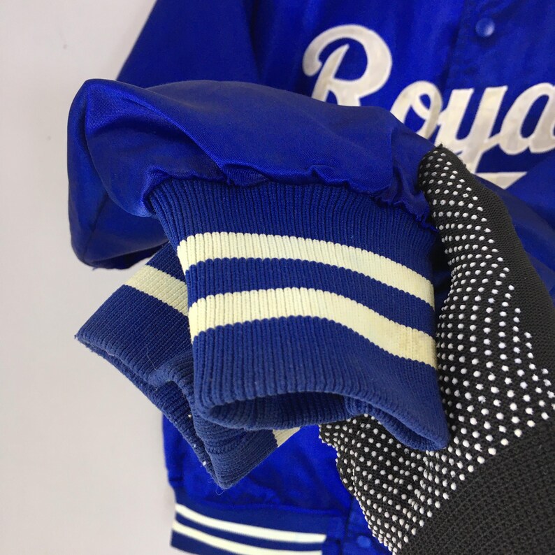 Pu&ograve; includere: Primo piano di una manica di giacca da baseball in raso blu. Il polsino della manica &egrave; blu con strisce bianche. La giacca ha la scritta "Royals" in bianco sul retro. La giacca &egrave; uno stile classico.