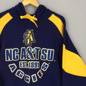 May include: Navy blue hoodie with yellow shoulder and hood panels. The front displays "NC A&T SU" and "EST. 1891" in white and blue, with "AGGIES" in an arc. A yellow logo is positioned above the text. The hoodie has a drawstring.