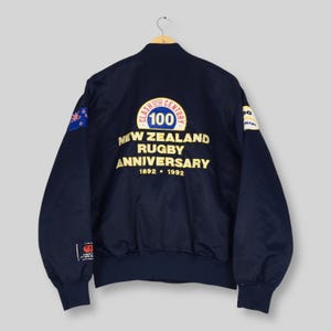 May include: Navy blue bomber jacket with gold lettering and patches. The jacket commemorates the New Zealand Rugby Anniversary, with text reading "Clash of the Century 100 New Zealand Rugby Anniversary 1892-1992". Features a New Zealand flag patch.