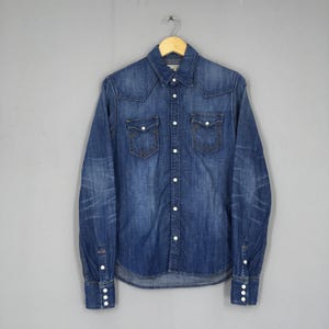May include: A blue denim shirt with a classic design, featuring two chest pockets and white buttons. The long-sleeved shirt is hanging on a wooden hanger against a gray background. The shirt has a faded, worn look.