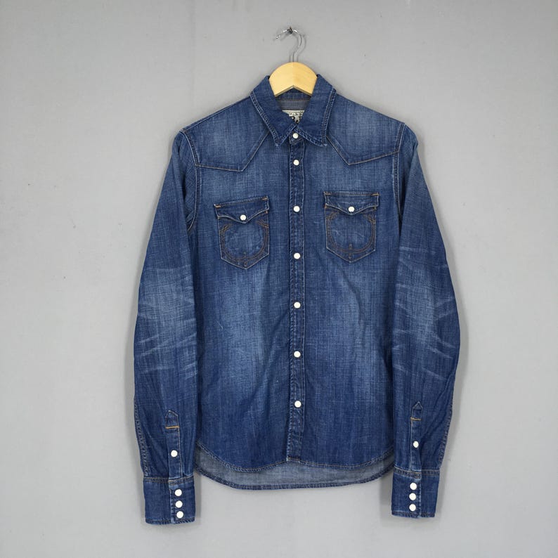 May include: A blue denim shirt with a classic design, featuring two chest pockets and white buttons. The long-sleeved shirt is hanging on a wooden hanger against a gray background. The shirt has a faded, worn look.