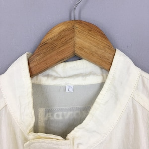 May include: A cream-colored collared jacket hanging on a wooden hanger. The jacket features a pointed collar, a visible size L tag at the inner neckline, and a partially obscured graphic print on the chest area. The fabric appears lightweight.