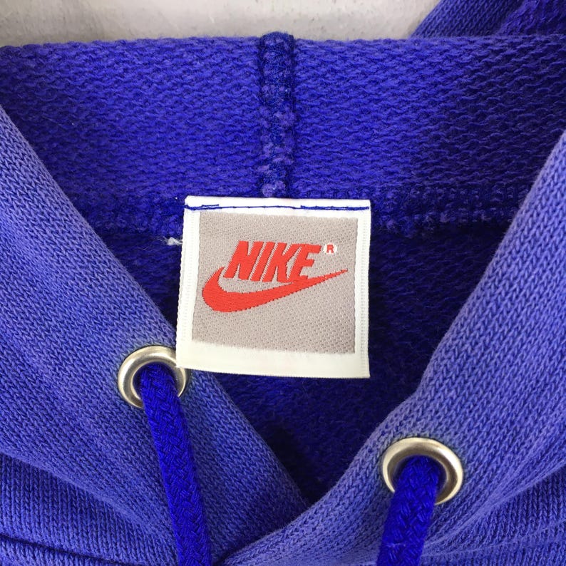May include: Close-up of a blue hoodie with a drawstring and metal eyelets. The Nike logo is displayed on a rectangular tag in the center. The logo is red, and the text is white.
