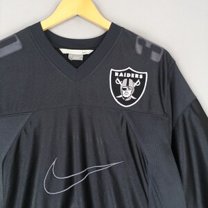 Puede incluir: Camiseta de f&uacute;tbol negra de los Raiders con cuello en V y el logo de Nike. La camiseta presenta el logotipo del equipo de los Raiders en el pecho y hombreras grises. La camiseta est&aacute; colgada en una percha de madera.