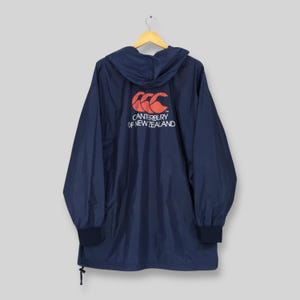 May include: A navy blue hooded windbreaker jacket featuring long sleeves with elastic cuffs and a drawstring hem. The back displays a red Canterbury logo above the white text CANTERBURY OF NEW ZEALAND, printed clearly on the center of the back.