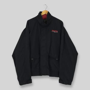 May include: Black Toyota motorsports jacket with a red lining. The jacket features a high collar, two front pockets, and the Toyota logo in red on the chest. It is displayed on a wooden hanger.