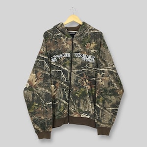 May include: A camouflage-patterned zip-up hoodie with the words "TRUE TIMBER CAMO" in white. The sweatshirt has brown trim on the cuffs, hem, and hood lining. The fabric features a woodland camouflage design, perfect for outdoor activities. The hoodie is hanging on a wooden hanger.
