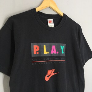 Puede incluir: Camiseta negra de Nike con la palabra "PLAY" en letras coloridas. Debajo, el texto "PARTICIPATE IN THE LIVES OF AMERICA'S YOUTH". El logo de Nike es rojo y naranja.