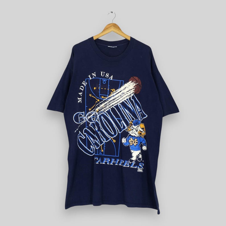 May include: Navy blue t-shirt featuring a basketball graphic with the words "Go Carolina" and "Tar Heels." The design includes a basketball, a basketball court diagram, and a cartoon mascot. The shirt is made in the USA.