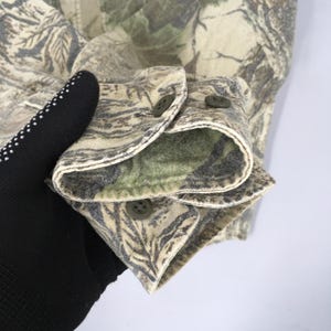 Puede incluir: Primer plano de una manga de camisa con estampado de camuflaje. La manga est&aacute; abotonada y presenta un forro interior verde. La tela tiene una mezcla de colores verdes, marrones y beige, con un dise&ntilde;o de hojas. Los botones son verdes.