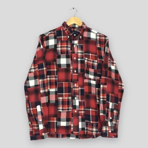Puede incluir: Una camisa abotonada de manga larga con un diseño de patchwork. La camisa presenta un patrón de cuadros rojos, negros, blancos y azul marino. La camisa está colgada en una percha de madera.