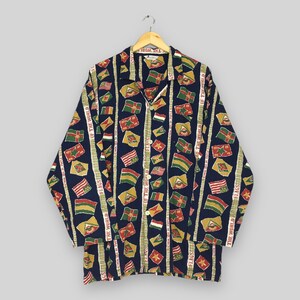 Puede incluir: Camisa de manga larga azul marino con un estampado repetido de banderas rojas, amarillas y verdes. Rayas verticales con el texto "THE WORLD FLAGS" recorren la camisa.
