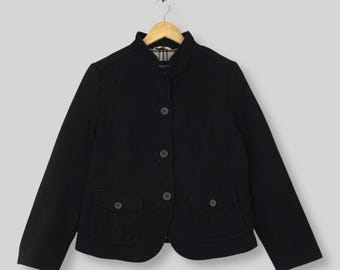 May include: A black jacket with a high collar, long sleeves, and a front button closure. The garment features two front pockets with buttoned flaps and a visible tan, black, and red plaid lining at the inner neckline, hanging on a wooden hanger.