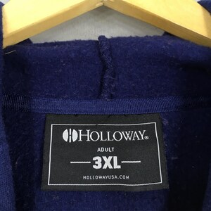May include: A navy blue hooded sweatshirt with a label displaying the brand name "HOLLOWAY" and the size "3XL". The label also states "ADULT" and "HOLLOWAYUSA.COM". The sweatshirt is suspended on a wooden hanger.