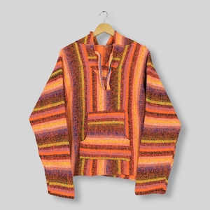 May include: A striped Baja hoodie in shades of orange, yellow, purple, and brown. The pullover features a hood, a front pocket, and a drawstring neckline. The garment is made of a woven fabric. The hoodie is hanging from a wooden hanger.