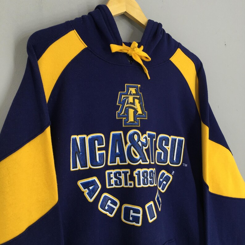 May include: Navy blue hooded sweatshirt with yellow panels on the sleeves and hood. The front displays "NCA & TSU" and "AGGIE" in white and blue lettering, with the established year "EST. 1891".