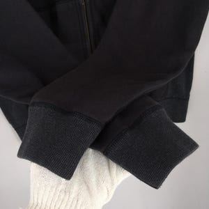 Pu&ograve; includere: Primo piano di una felpa con cappuccio nera con cerniera. I polsini sono di una tonalit&agrave; di nero pi&ugrave; scura con un motivo a maglia strutturato. La cerniera &egrave; visibile e le maniche sono piegate l'una sull'altra.