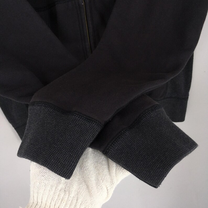 Pu&ograve; includere: Primo piano di una felpa con cappuccio nera con cerniera. I polsini sono di una tonalit&agrave; di nero pi&ugrave; scura con un motivo a maglia strutturato. La cerniera &egrave; visibile e le maniche sono piegate l'una sull'altra.