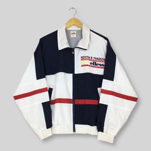 May include: A vintage white, navy, and red windbreaker jacket with the text "NOVITA' E TRADIZIO" and "ellesse" printed on the chest. The jacket has a zip-up front, a white collar, and red stripes across the front.