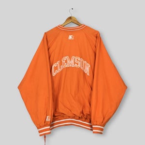 Puede incluir: Chaqueta naranja de Clemson con detalles blancos. La palabra "CLEMSON" está impresa en letras blancas grandes en la espalda. El logotipo de Starter es visible en la parte superior de la espalda y en la manga. La chaqueta tiene un cordón en la cintura y rayas blancas en los puños y el dobladillo.