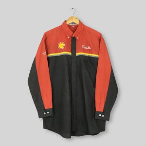 Puede incluir: Camisa abotonada de manga larga con sección superior roja y sección inferior negra. La camisa presenta el logotipo de Shell, la palabra "HELIX" y otro texto. La camisa está colgada de una percha de madera.
