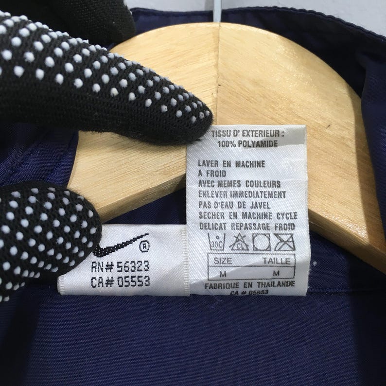 Pu&ograve; includere: Primo piano di un'etichetta di abbigliamento blu navy con testo bianco e un logo Nike. L'etichetta include le istruzioni di lavaggio, la taglia M e il testo "FABRIQUE EN THAILANDE". Il capo &egrave; realizzato in 100% poliammide.