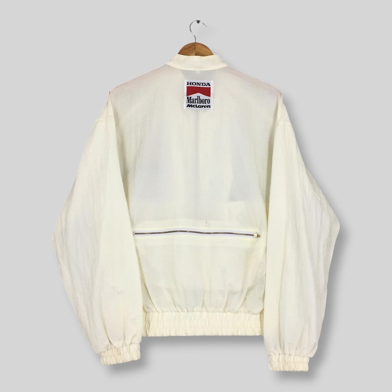 May include: A white long-sleeve jacket with a high collar and elasticized cuffs and hem. The back features a rectangular patch with the text Honda, Marlboro, and McLaren in red, white, and black. A horizontal zippered pocket is located below the patch.