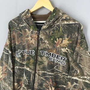May include: A camouflage zip-up hoodie with the text "TRUE TIMBER CAMO" in white. The hoodie features a hood, a full-length zipper, and a brown interior lining. The camouflage pattern includes shades of brown, green, and tan.