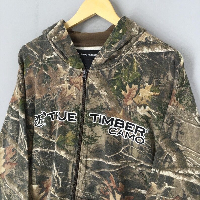 May include: A camouflage zip-up hoodie with the text "TRUE TIMBER CAMO" in white. The hoodie features a hood, a full-length zipper, and a brown interior lining. The camouflage pattern includes shades of brown, green, and tan.