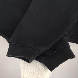 May include: Close-up of a black sweatshirt sleeve. The sleeve is made of a thick, dark fabric with a ribbed cuff. The cuff is a darker shade of black than the rest of the sleeve. The image is well-lit, showing the texture of the fabric.