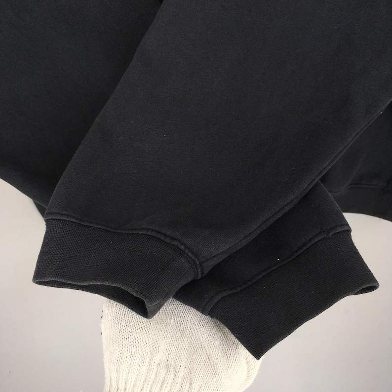 May include: Close-up of a black sweatshirt sleeve. The sleeve is made of a thick, dark fabric with a ribbed cuff. The cuff is a darker shade of black than the rest of the sleeve. The image is well-lit, showing the texture of the fabric.