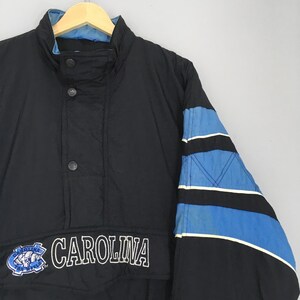 May include: Black jacket with blue and white accents. The jacket features the word "CAROLINA" in white lettering, along with a logo. The sleeves have a blue and black design. The jacket has a stand-up collar and three buttons.