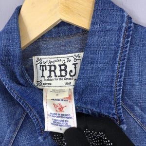 May include: Close-up of a blue denim jacket with a collar and a wooden hanger. The jacket has a label that reads "TRBJ Fashion for the Senses" and "Brand Jeans". The jacket is size S and made in Mexico.