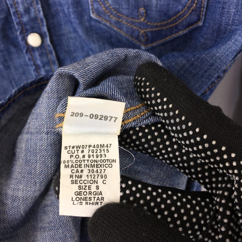 May include: Close-up of a blue denim shirt with a white tag attached. The tag displays the number "209-092977" and other text including "SIZE S" and "LONESTAR L/S SHIRT". The shirt has a button closure.