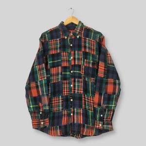 Puede incluir: Una camisa abotonada de manga larga con un diseño de cuadros patchwork. La camisa presenta una mezcla de patrones de cuadros rojos, verdes, azul marino y negros. Cuelga de una percha de madera sobre un fondo gris claro.