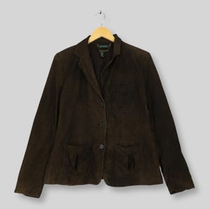 May include: A dark brown suede blazer featuring a notched lapel, a three-button front closure, one chest pocket, and two lower patch pockets. The interior label reads Lauren. The garment is displayed on a wooden hanger against a neutral background.